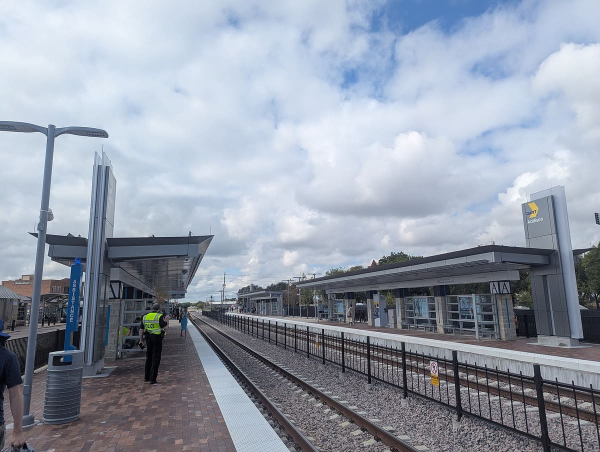 The two Silver Line platforms at Addison Station with steel canopies, a DART Addison sign tower, and brick paving under a partly cloudy sky