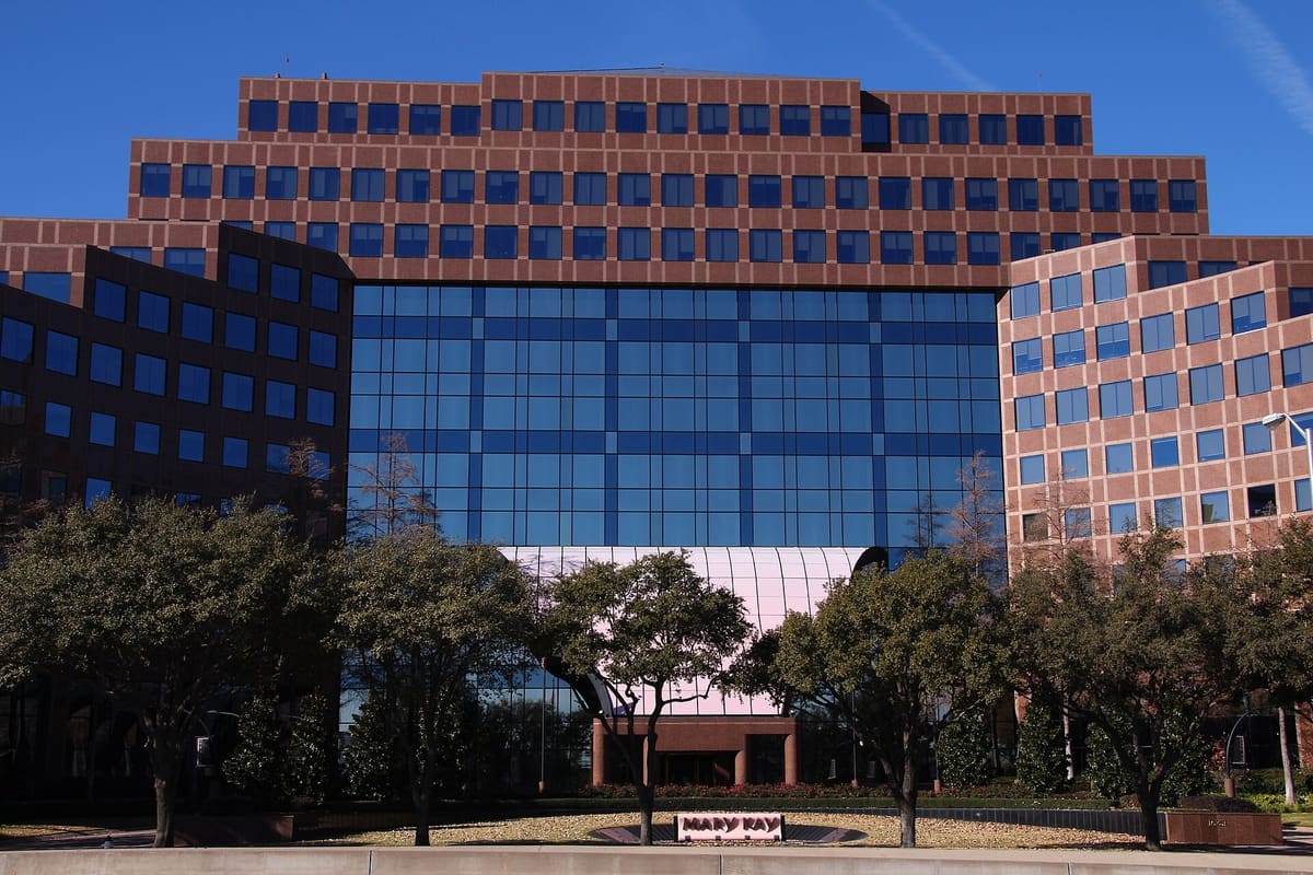 The Mary Kay headquarters on Dallas Parkway in Addison, a stepped red granite office building with a tall blue glass atrium behind a row of live oaks