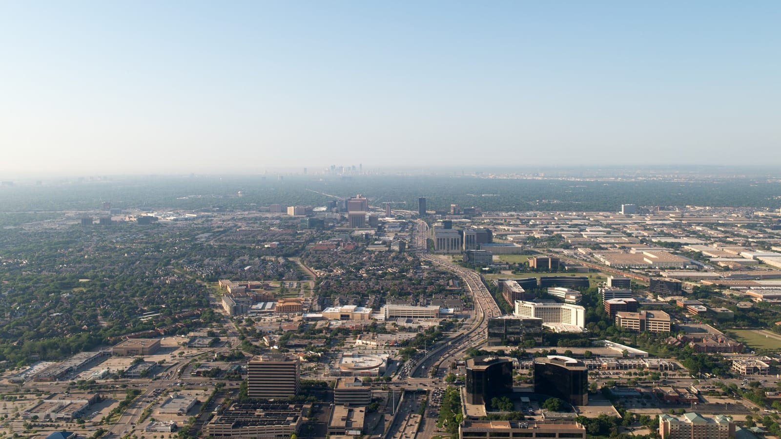 Aerial view over Addison, Texas, looking south along the Dallas North Tollway past office towers and hotels toward the downtown Dallas skyline on the horizon