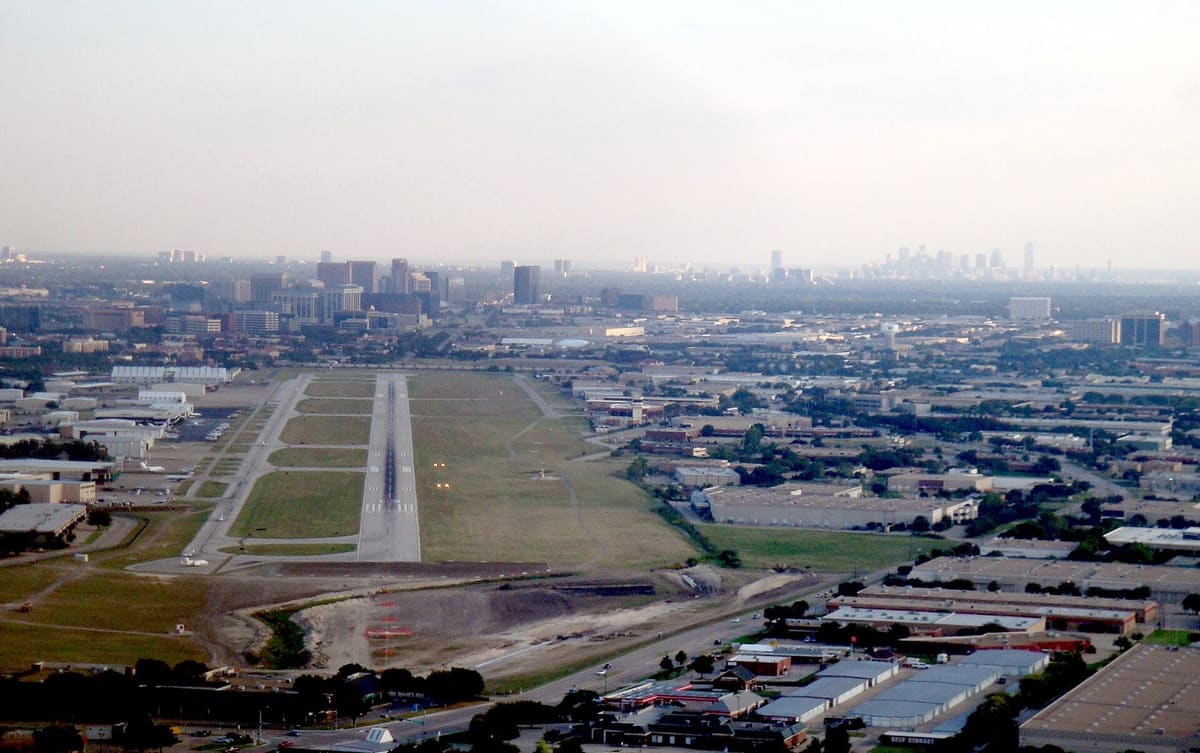 Aerial view of the Addison Airport runway stretching south between hangars and business parks, with the Tollway office towers and downtown Dallas on the horizon
