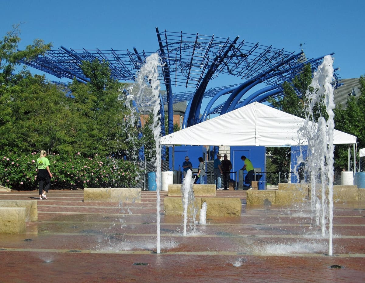 Ground fountains on the brick plaza at Addison Circle with the blue steel petals of the Blueprints sculpture rising behind the trees
