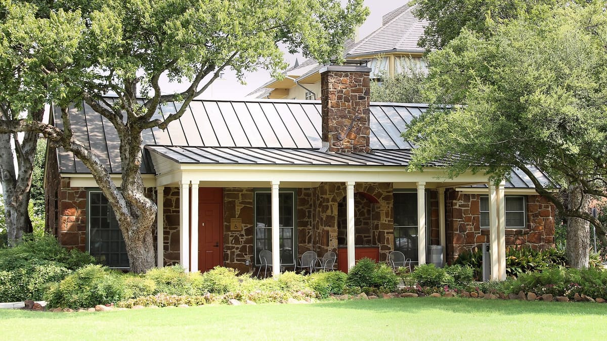 The Stone Cottage in Addison, Texas, a one-story WPA building with red-brown rock walls, a standing seam metal roof, a stone chimney, and a white columned porch under shade trees