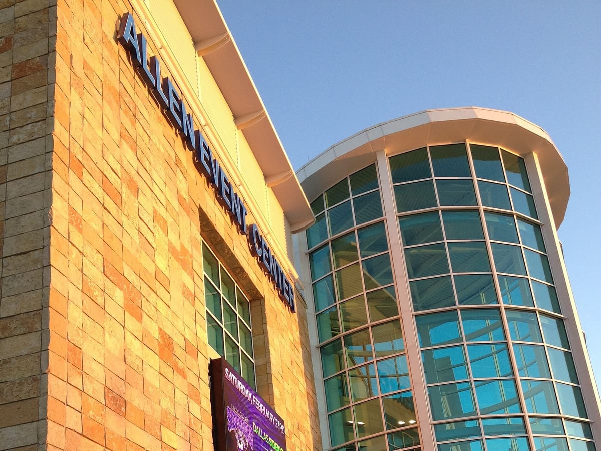 The stone and glass facade of the Allen Event Center in Allen, Texas, with its name in raised letters and a curved glass tower lit by evening sun