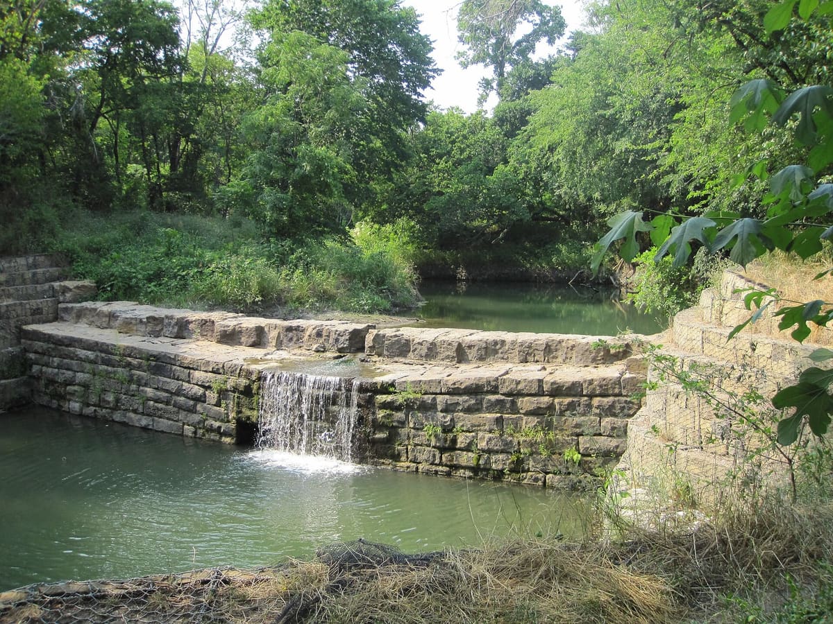 Water spilling over the 1874 limestone block dam on Cottonwood Creek in Allen, Texas, surrounded by green trees