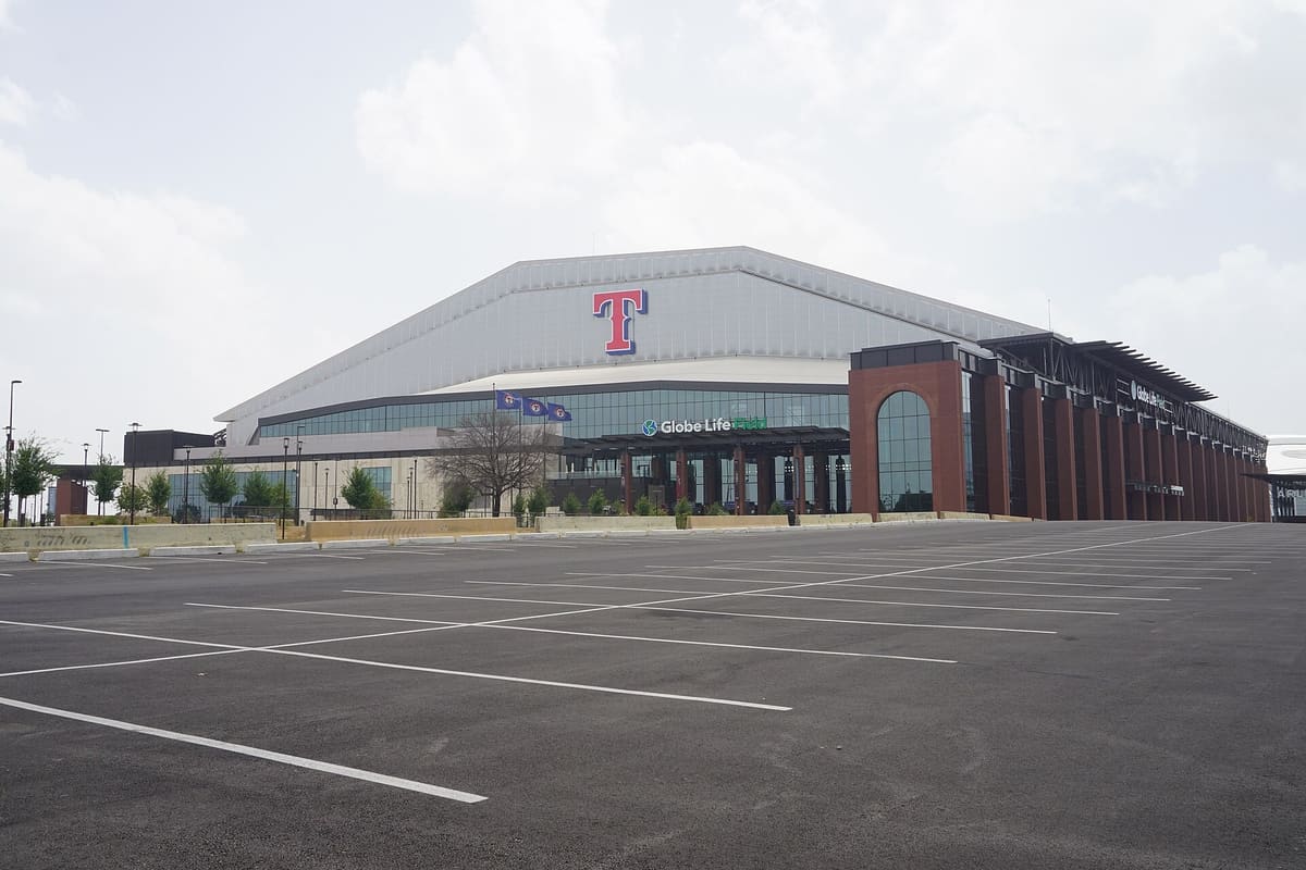 Globe Life Field in Arlington, a brick and glass ballpark with a pale arched roof and a red T logo, seen across an empty parking lot