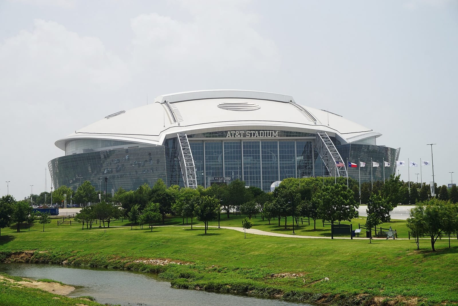 AT&T Stadium in Arlington, its arched white roof and glass end wall rising beyond a creek and a park lawn with young trees
