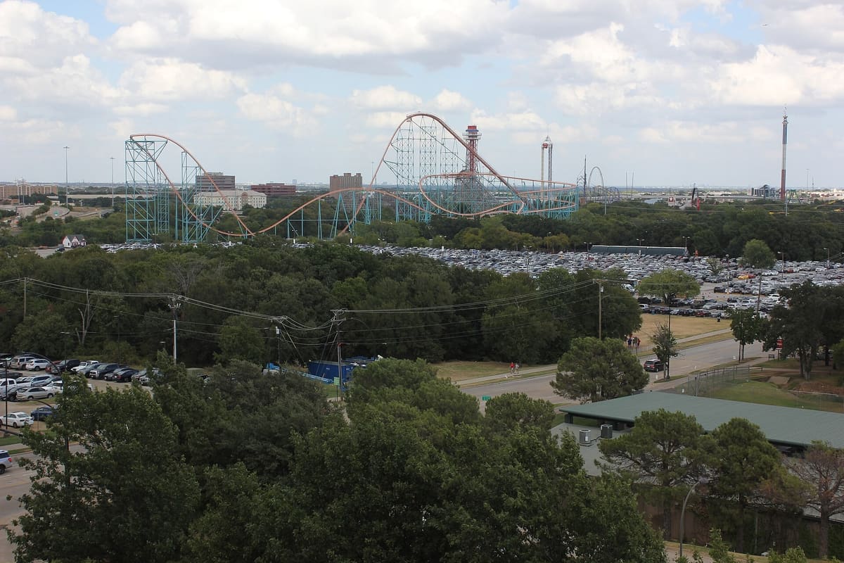 Six Flags Over Texas seen from above, with roller coaster tracks and an observation tower rising over a tree line and large parking fields