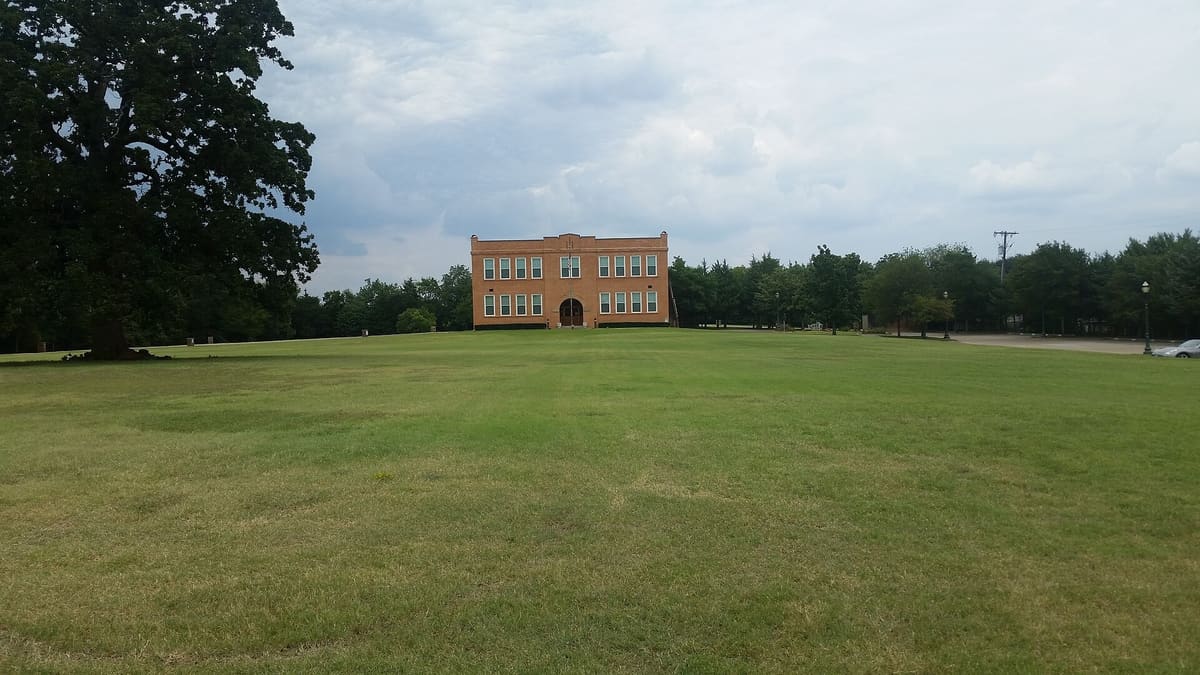 The Old Bedford School seen across a wide green lawn, with a large oak tree at left and a row of trees behind the two-story brick building
