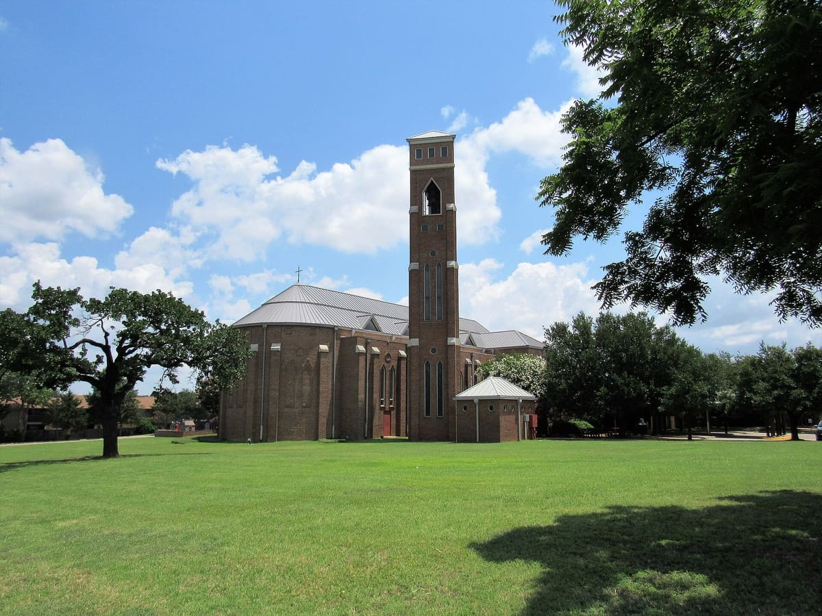 St. Vincent's Cathedral in Bedford, Texas, a brick church with a standing seam metal roof and a tall square brick bell tower beside a wide lawn and an oak