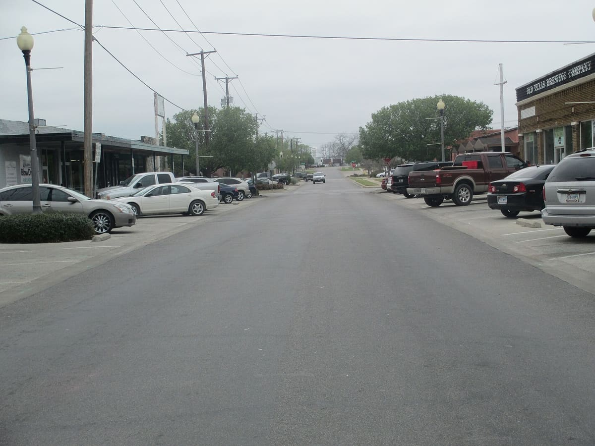 A street in Old Town Burleson with a low storefront row under a flat metal canopy on the left and brick commercial buildings on the right, cars parked nose in on both sides