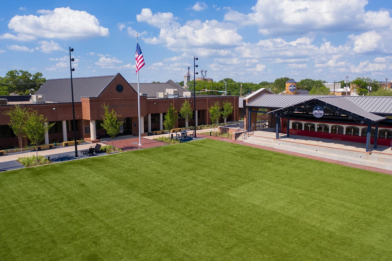 Burleson City Hall's red brick front and the lawn of Mayor Vera Calvin Plaza, with a red interurban parlor car under a covered pavilion on the right