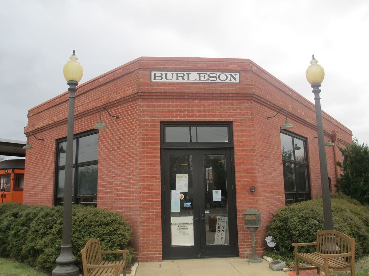 The restored brick interurban depot in Burleson with a BURLESON sign over glass double doors, globe street lamps, wooden benches, and an orange rail car at the left edge