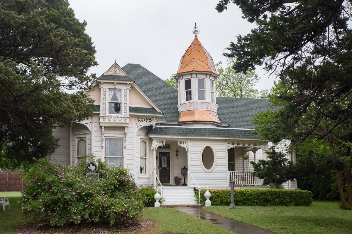 The Renfro-Clark House in Burleson, a white Victorian frame house with a copper-clad turret, scrollwork porch brackets, an oval window, and a Texas historical marker among blooming shrubs