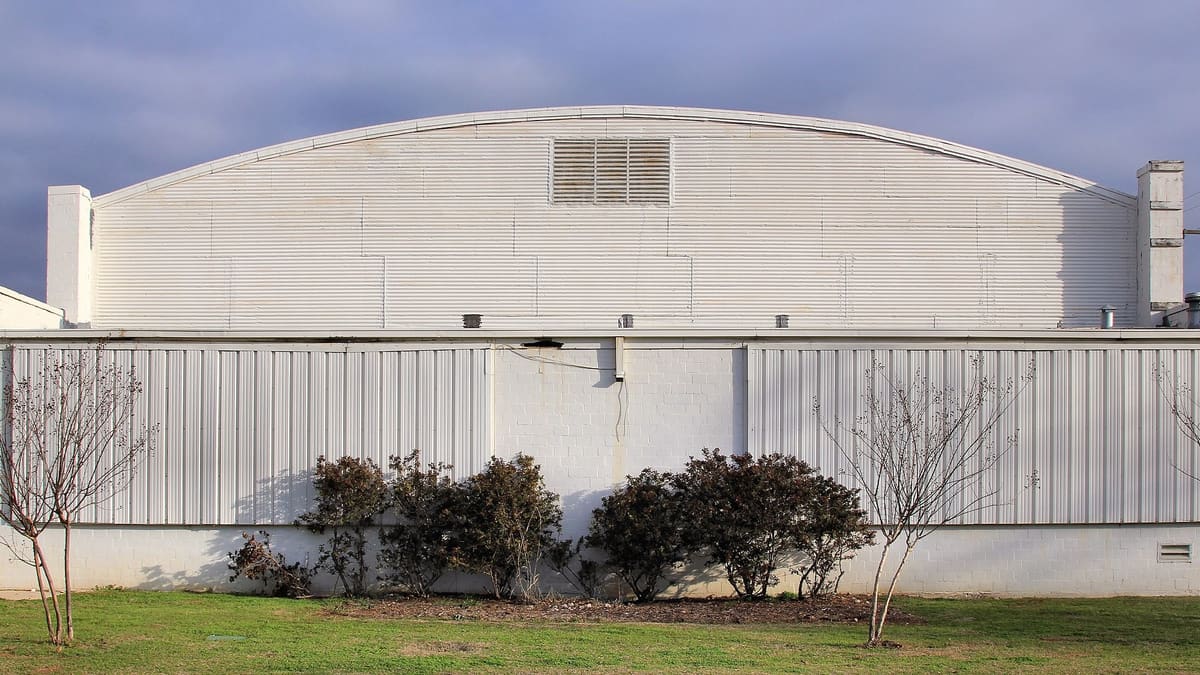 The south side of the 1936 Burleson Gymnasium with a white barrel roof, corrugated metal gable, painted block walls, and long metal panels along the upper wall