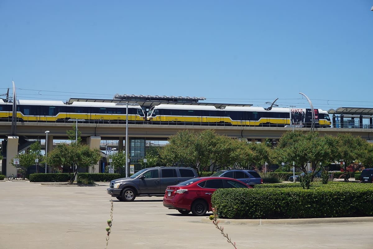 Two yellow and white DART light rail trains on the elevated Green Line platform at Downtown Carrollton Station, above a landscaped parking lot