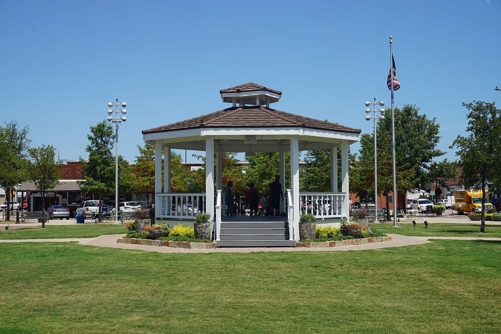 The white gazebo at the center of the Carrollton Town Square, with brick storefronts and a flagpole behind it on a clear summer day