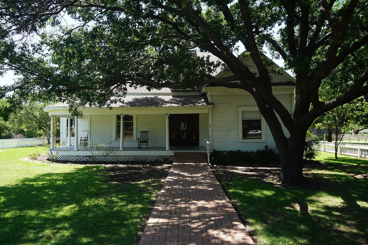 The white frame A.W. Perry Homestead Museum in Carrollton with a wraparound porch and rocking chairs, shaded by a large oak, with a brick walk leading to the front door