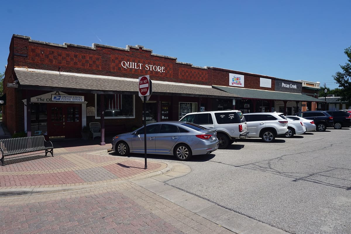 A row of one-story red brick storefronts on West Main Street at the Carrollton square, with decorative brickwork, a shingled canopy, and cars parked out front