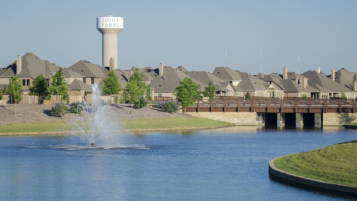 Two-story brick and stone homes in Light Farms in Celina, Texas, across a pond with a fountain and a wood pedestrian bridge, with the Light Farms water tower behind