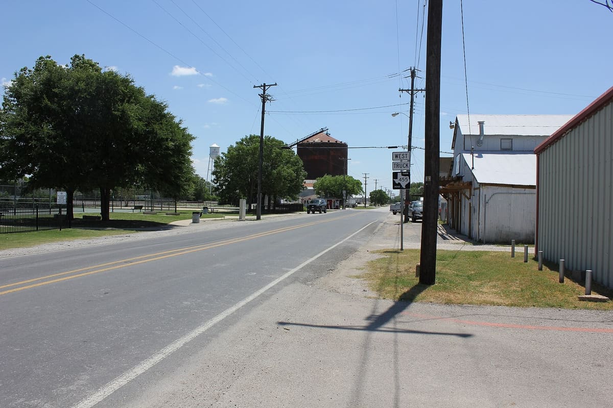 North Louisiana Drive near downtown Celina, Texas, with white metal-sided buildings, a large rusted grain tank, and the Celina water tower in the distance