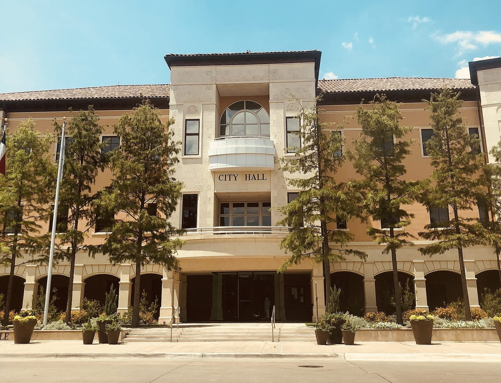 Colleyville City Hall at 100 Main Street, a three-story building with a clay tile roof, a cast stone entry tower, and an arched loggia behind a row of young trees