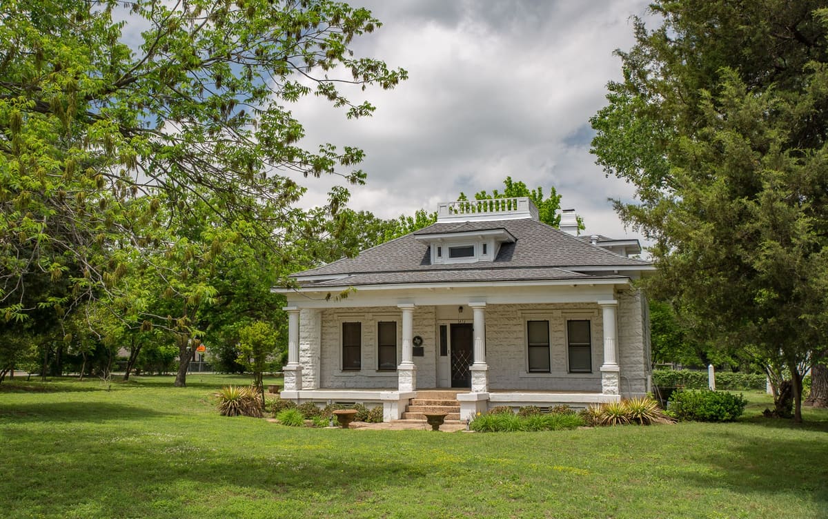The Bidault House in Colleyville, a white single-story home of molded concrete block with a hipped gray roof, a columned front porch, and a Texas historical medallion beside the door