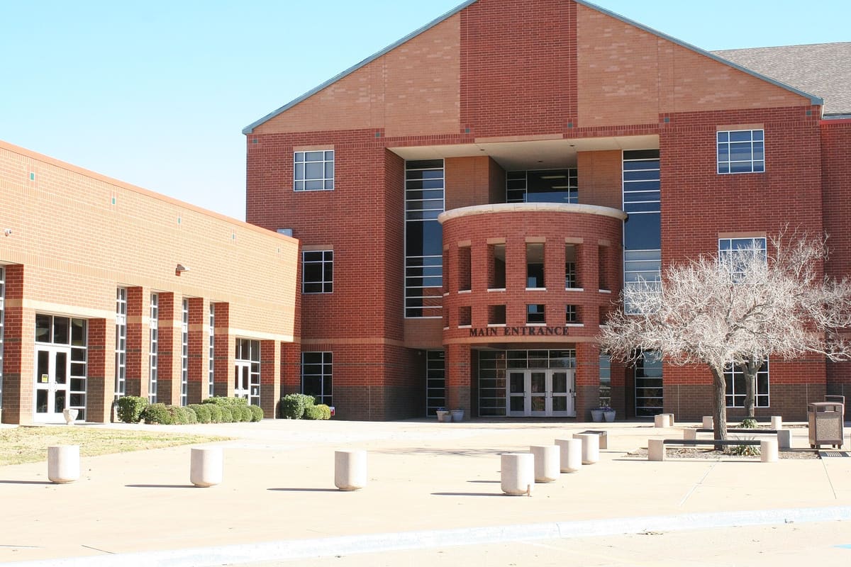 The main entrance of Colleyville Heritage High School, a red brick building with a rounded brick entry tower, tall window bands, and concrete bollards across the plaza