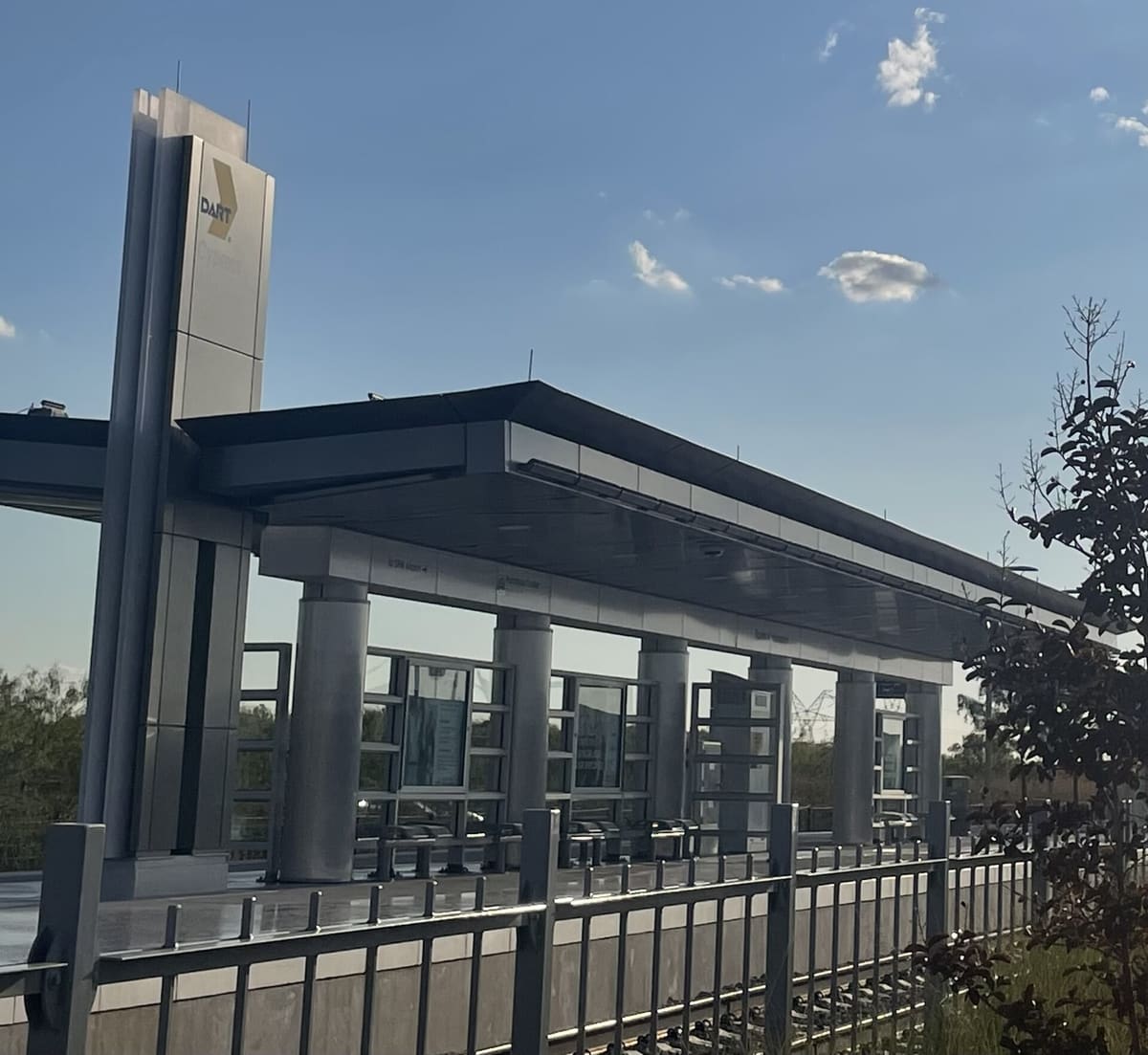 Cypress Waters station on DART's Silver Line, a steel platform canopy beside a tall DART pylon sign under a blue sky with a few clouds