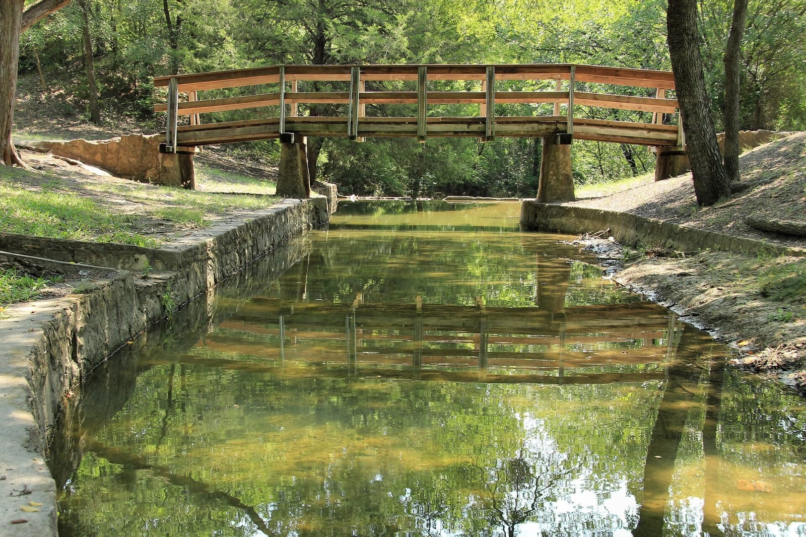 A wooden footbridge on stone piers over the rock-lined spring channel at Grapevine Springs Park in Coppell, Texas, reflected in still green water under shade trees