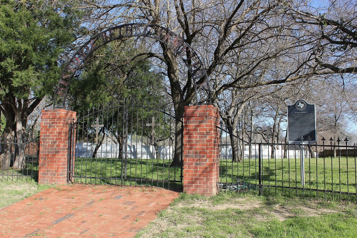 The brick and iron entrance gate of Historic Bethel Cemetery in Coppell, Texas, with an arched sign, two crosses on the gate, and a Texas historical marker beside it