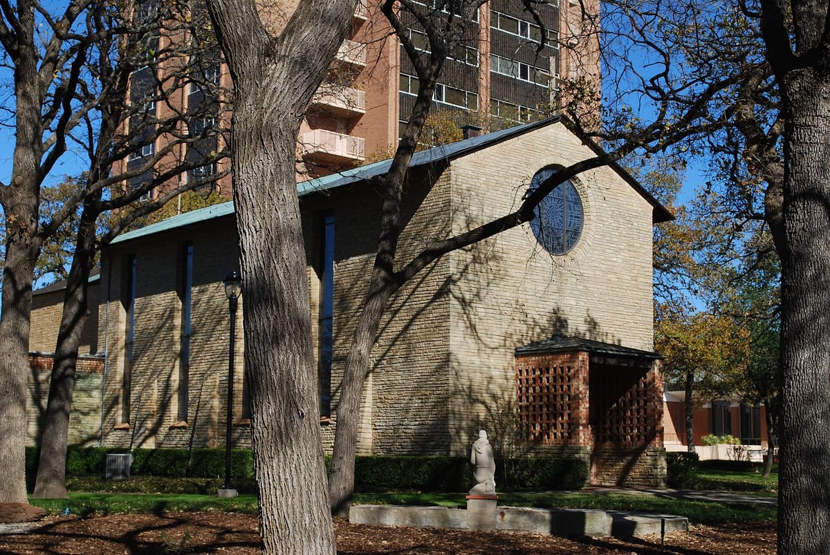 The Little Chapel-in-the-Woods at Texas Woman's University, a pale brick chapel with a round stained glass window and a brick lattice entry porch under oak trees