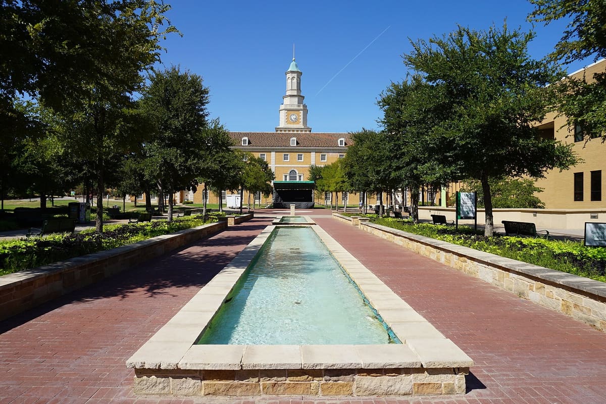 The Hurley Administration Building at the University of North Texas, a buff brick building with a white clock tower, behind a long reflecting pool and brick plaza