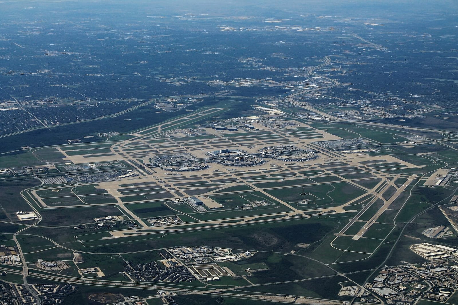 Aerial view of DFW International Airport from an arriving flight, with its runways, terminal loops, and the freeways and neighborhoods around its edges