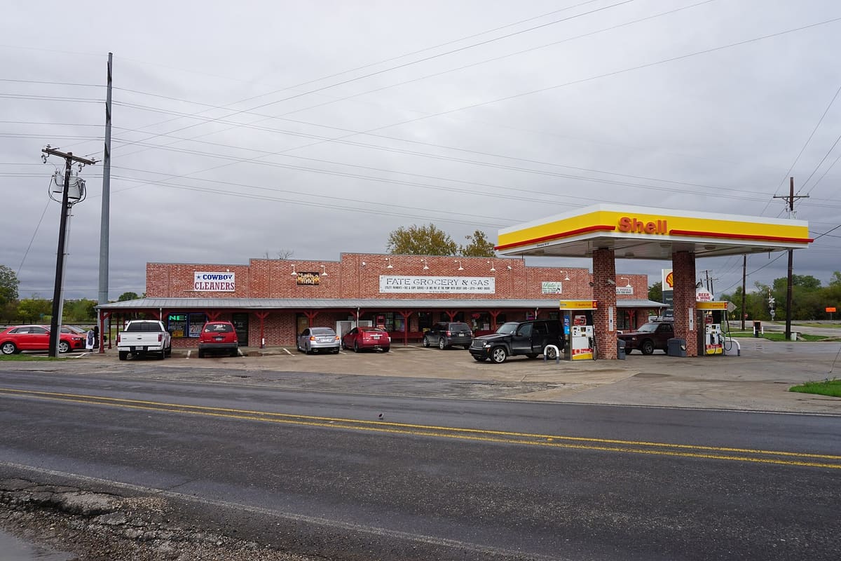 Fate Grocery and Gas, a one-story red brick retail strip with a metal-roofed covered walkway and a fuel canopy on West Fate Main Place in Fate, Texas