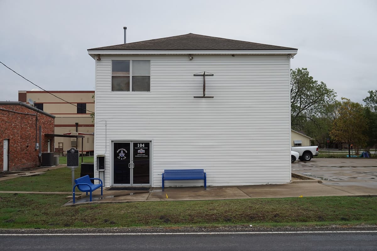 The two-story white sided Fate Lodge No. 802 hall at 104 South W.E. Crawford in Fate, Texas, signed as the City of Fate Community Center, with a state historical marker and blue benches