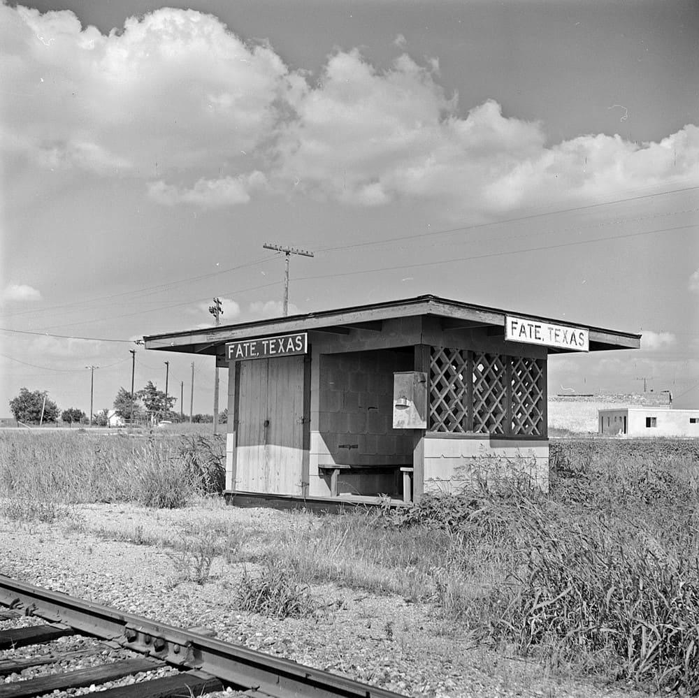 The small wooden M-K-T Railroad flag stop shelter at Fate, Texas, in June 1962, with Fate, Texas signs on both ends, lattice panels, and a bench beside the tracks