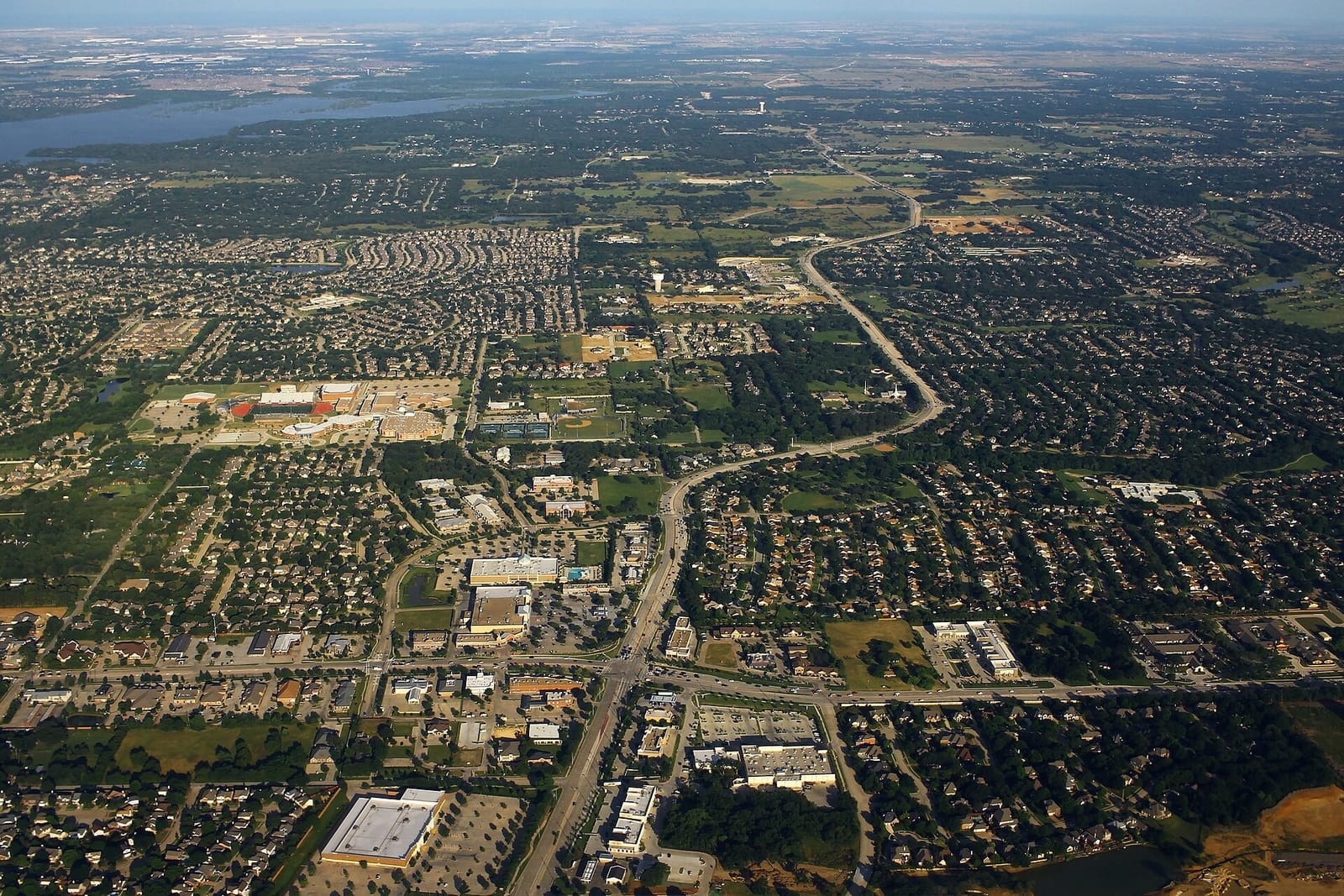 Aerial view of Flower Mound, Texas, with shopping centers along a divided road, neighborhoods under heavy tree cover, and lake water on the horizon