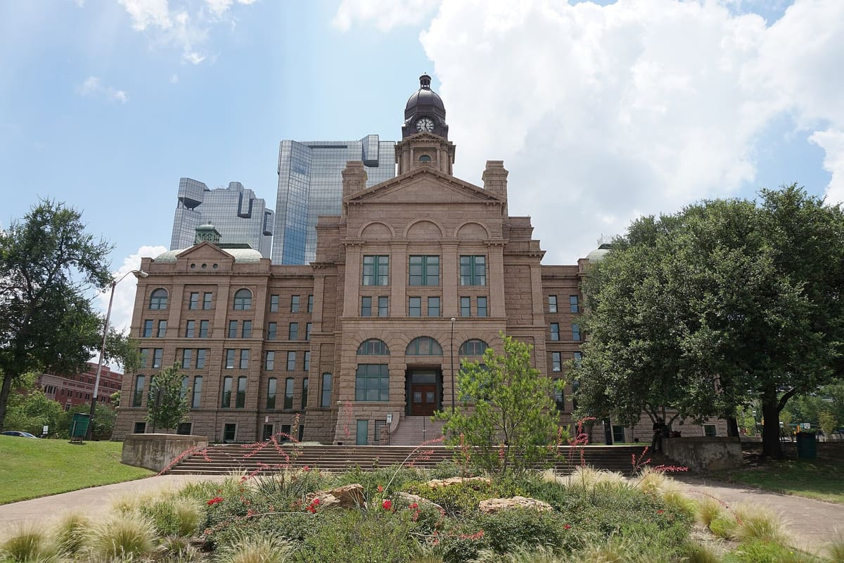 The pink granite Tarrant County Courthouse with its clock tower, framed by live oaks, with glass downtown towers rising behind it