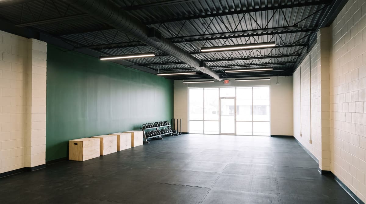 Fitness studio inside a converted 1960s concrete block building with painted block walls, black rubber flooring, exposed steel roof joists, and a new glass storefront