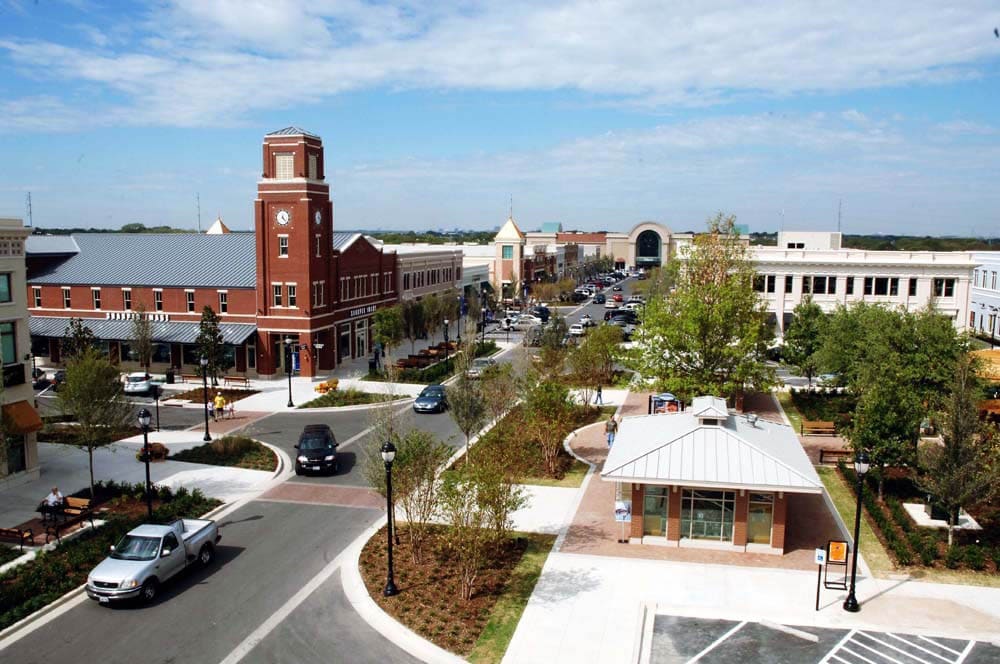 Aerial view west along a main street at Firewheel Town Center in Garland, with a brick clock tower, storefronts, young street trees, and a small pavilion