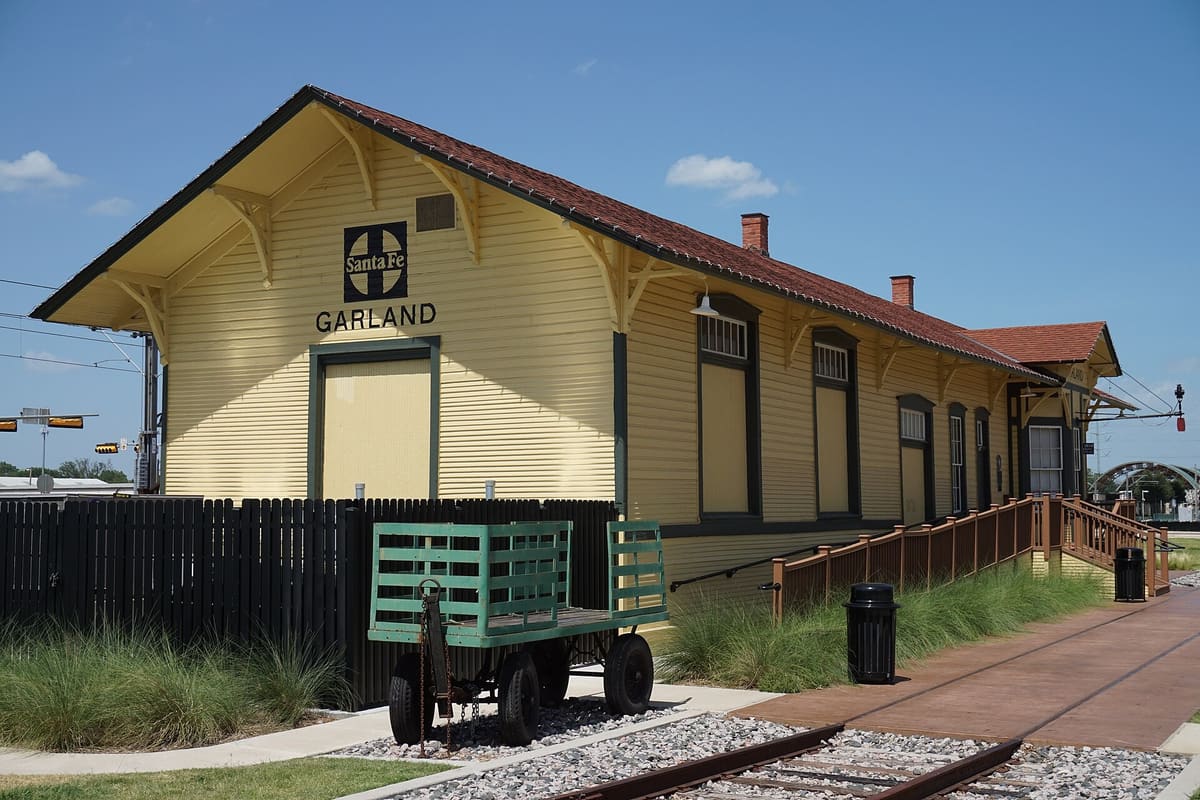 The yellow wood-frame 1901 Santa Fe depot in Garland, Texas, with a dark red roof, Santa Fe and Garland signs on the gable, and a green baggage cart beside the track