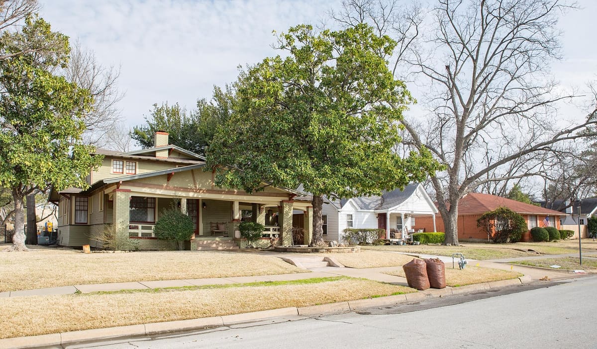 A Craftsman bungalow with a deep front porch and brick piers on South 11th Street in Garland's Travis College Hill Historic District, beside a white cottage and a brick ranch house