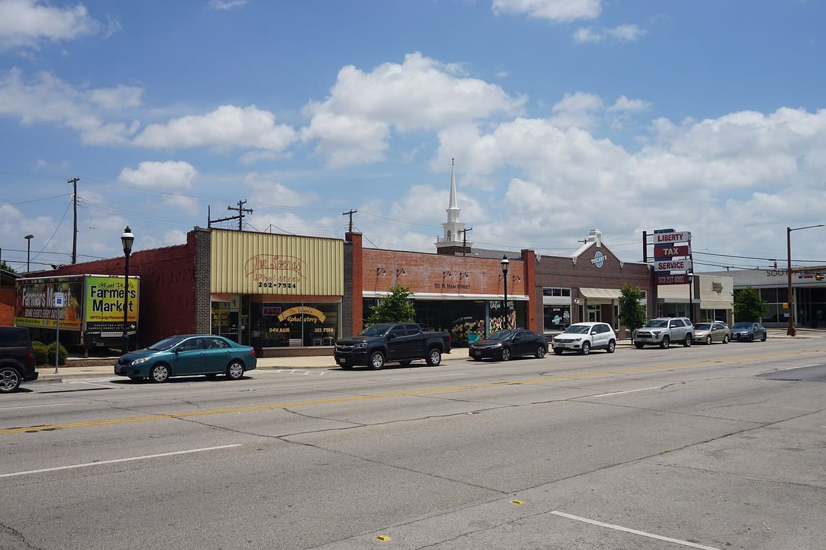 West Main Street in downtown Grand Prairie, a row of older one-story red brick storefronts with awnings and a white church steeple rising behind them
