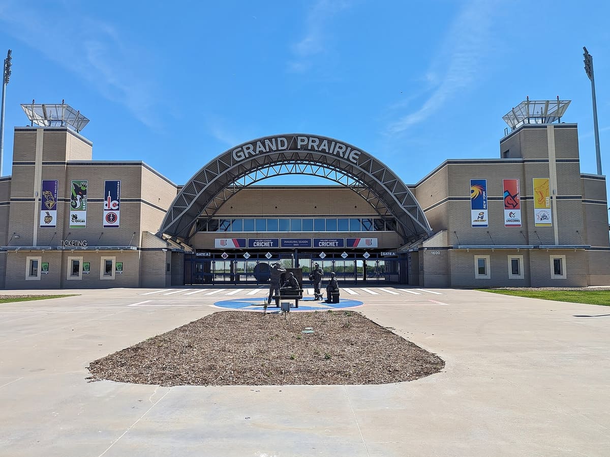 Grand Prairie Stadium on Lone Star Parkway, a tan masonry stadium entrance under a steel arch reading Grand Prairie, with team banners on two towers and a bronze statue group on the plaza
