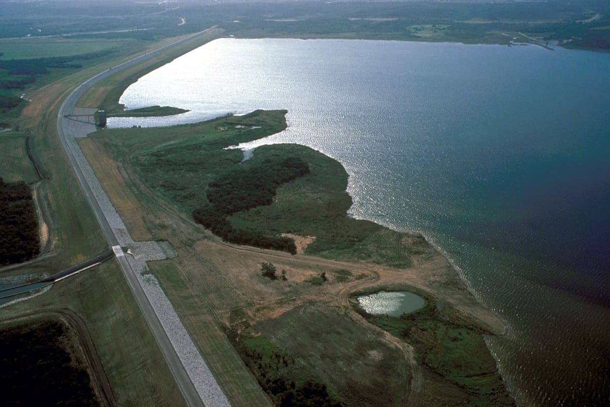 Aerial view of Joe Pool Lake and its earthen dam, with a road along the dam crest, green shoreline, and open water reaching to the horizon