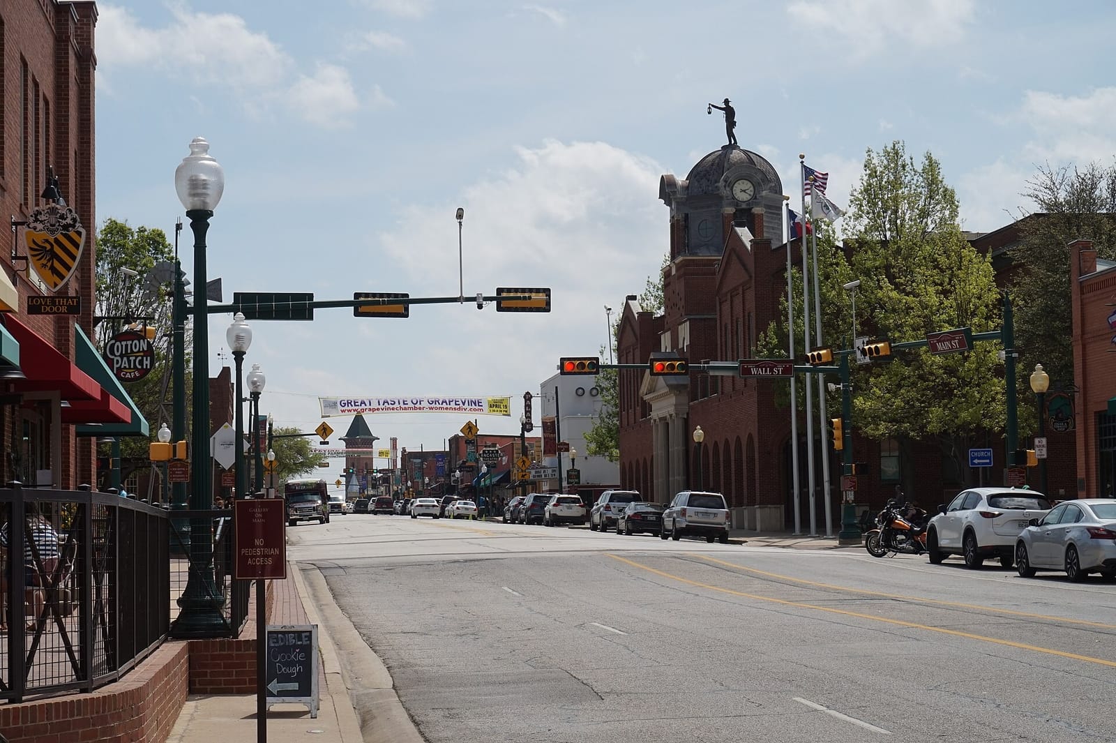 Main Street in historic downtown Grapevine, Texas, looking south past Wall Street, with red brick storefronts, the Palace Theatre sign, and the City Hall clock tower topped by the Nightwatchman statue