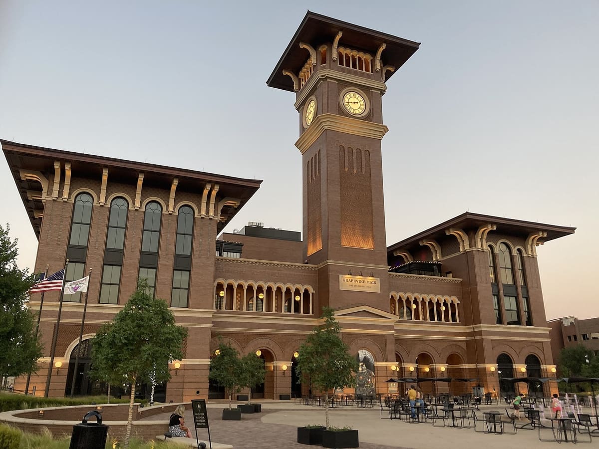 Grapevine Main Station at dusk, a brick building with arched windows and cream stone trim beside a tall brick clock tower, with tables on the plaza in front