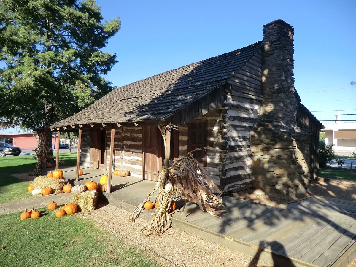 The Torian Log Cabin in Grapevine, a hand-hewn log house with white chinking, a stone chimney, wood shingle roof, and a shed porch decorated with pumpkins