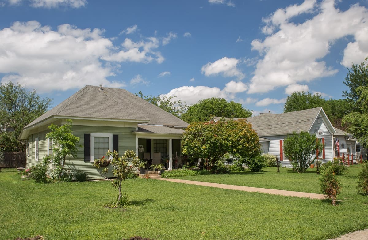 Two wood-frame houses in Grapevine's Original Town Residential Historic District, a sage green hipped-roof cottage with a recessed porch and a pale blue gabled house beside it