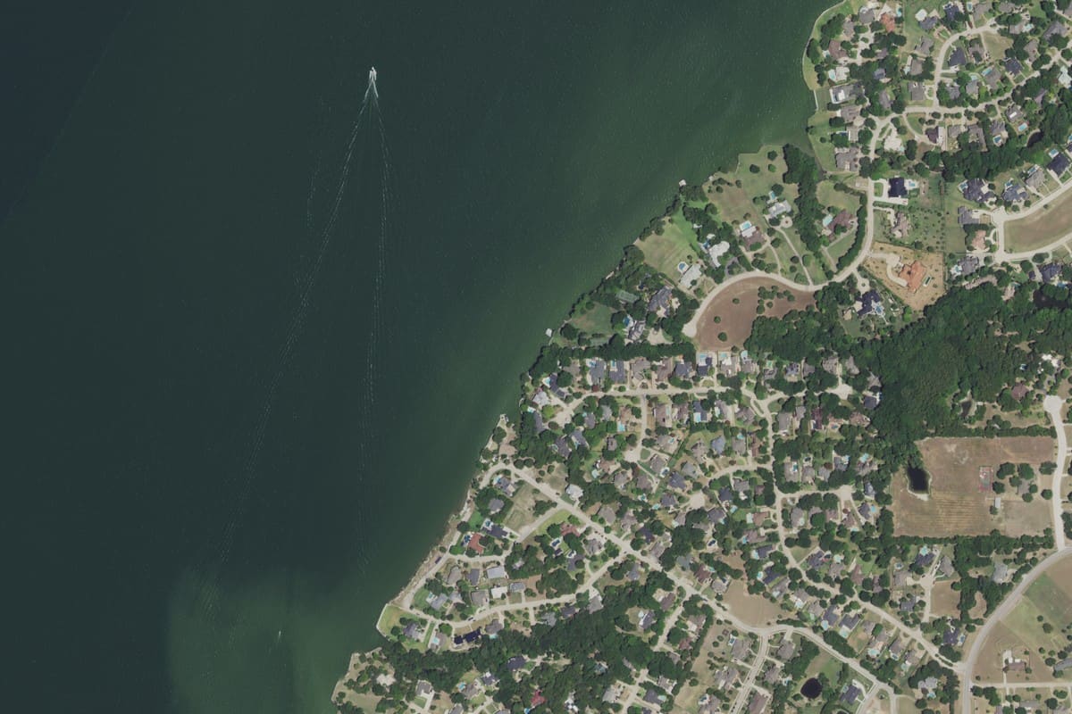 Aerial view of large lakefront home lots along Heath's Lake Ray Hubbard shoreline, with wooded yards, pools, and curving streets beside open water and a passing boat